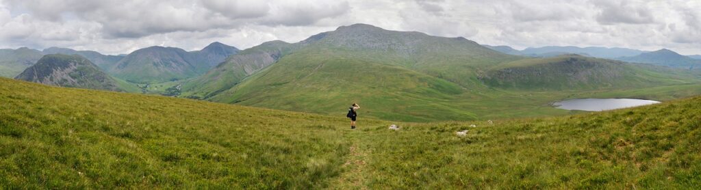 A woman in black back facing the camera in a vast green landscape of scafell pike with a lake on right in the mountains. 